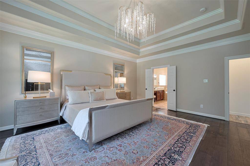 This elegant primary bedroom features a tray ceiling with a stunning new chandelier, a large bed with plush pillows, and matching side tables with lamps. The room is adorned with a stylish area rug and has an en-suite bathroom, offering a sophisticated and serene atmosphere.