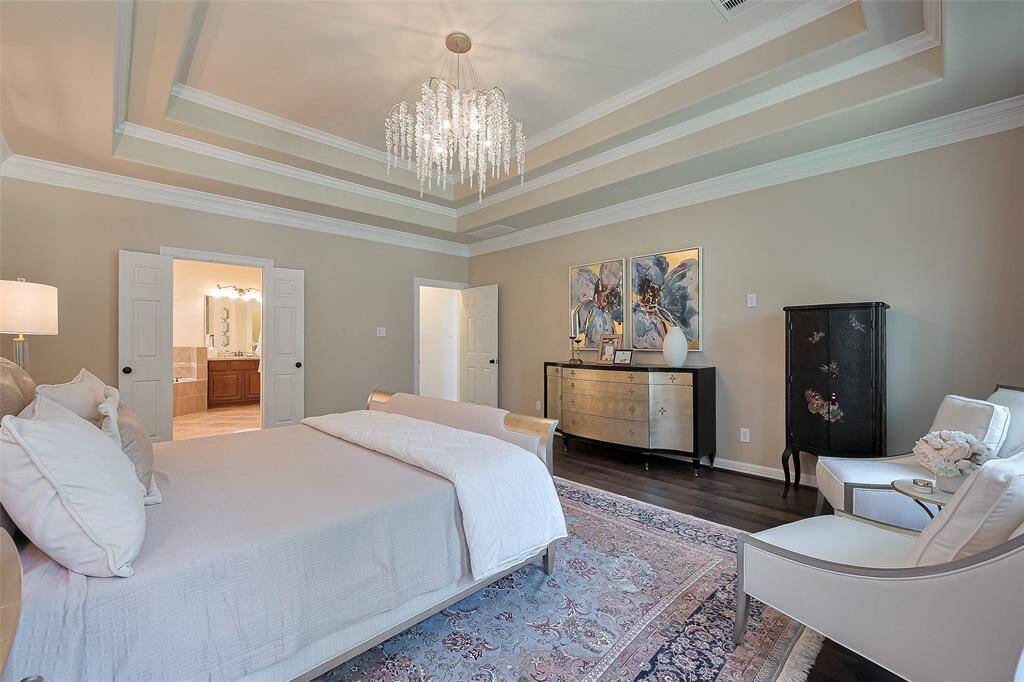This elegant primary bedroom features a luxurious tray ceiling with a chandelier, soft neutral walls, and rich wood flooring. It includes a comfortable seating area, stylish furniture, and a spacious layout that connects to a well-appointed en-suite bathroom.