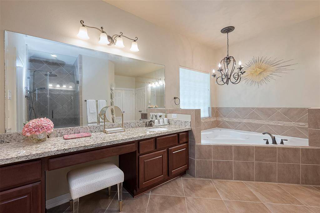 This spacious primary bathroom features a luxurious soaking tub, a large granite vanity with dual sinks, elegant lighting fixtures, and a separate glass-enclosed shower. The warm tile flooring and stylish decor create a relaxing atmosphere.