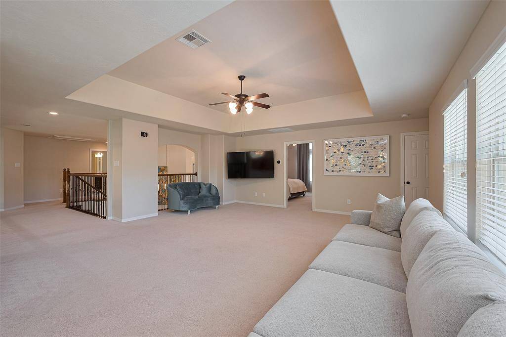 This spacious and bright second floor living area features neutral tones, ample natural light from large windows, and an open layout. It includes a ceiling fan, wall-mounted TV, and access to an adjacent room, offering a versatile space for relaxation or entertainment.