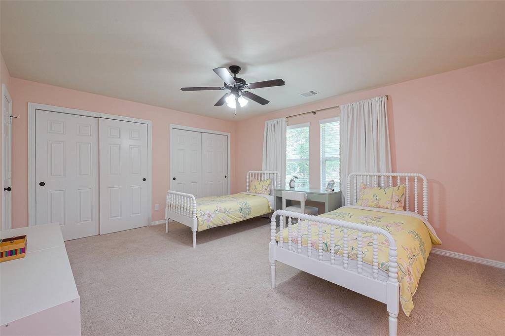 This spacious bedroom features soft pink walls, two twin beds with floral bedding, large windows with white curtains, ample closet space, and a ceiling fan, creating a cozy and inviting atmosphere.