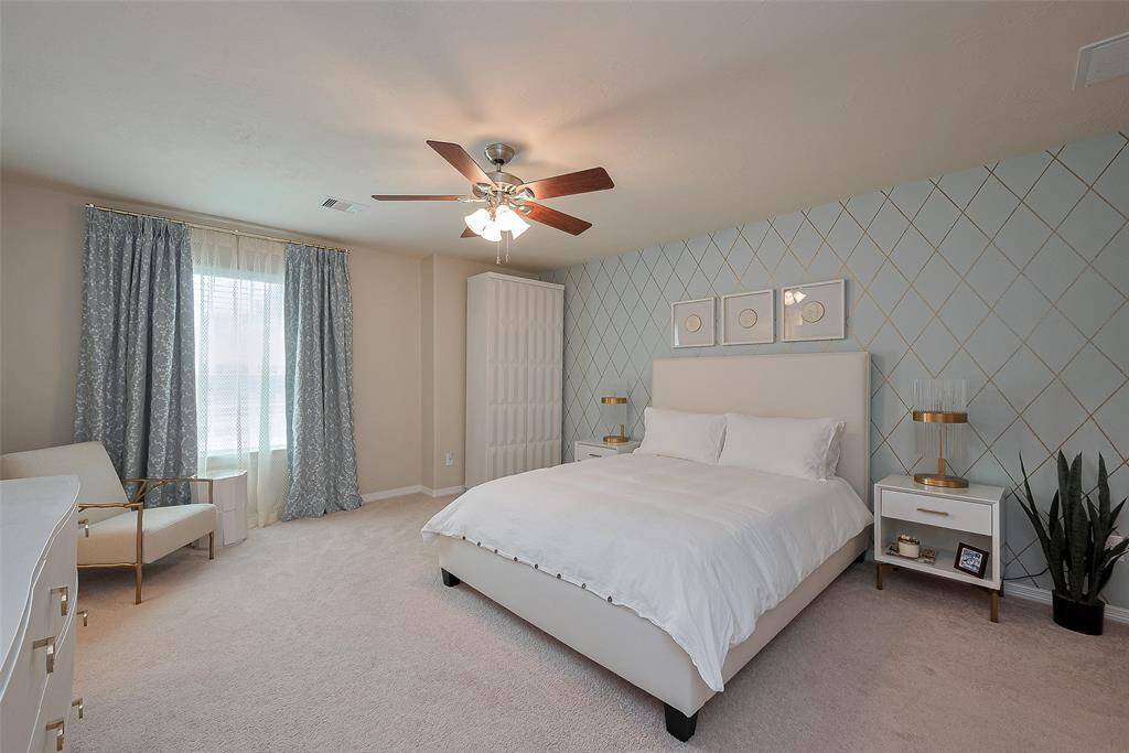 Spacious and bright bedroom with a modern geometric accent wall, ceiling fan, and soft carpeting. Features a comfortable bed, stylish side tables, and ample natural light from large windows with elegant drapes.
