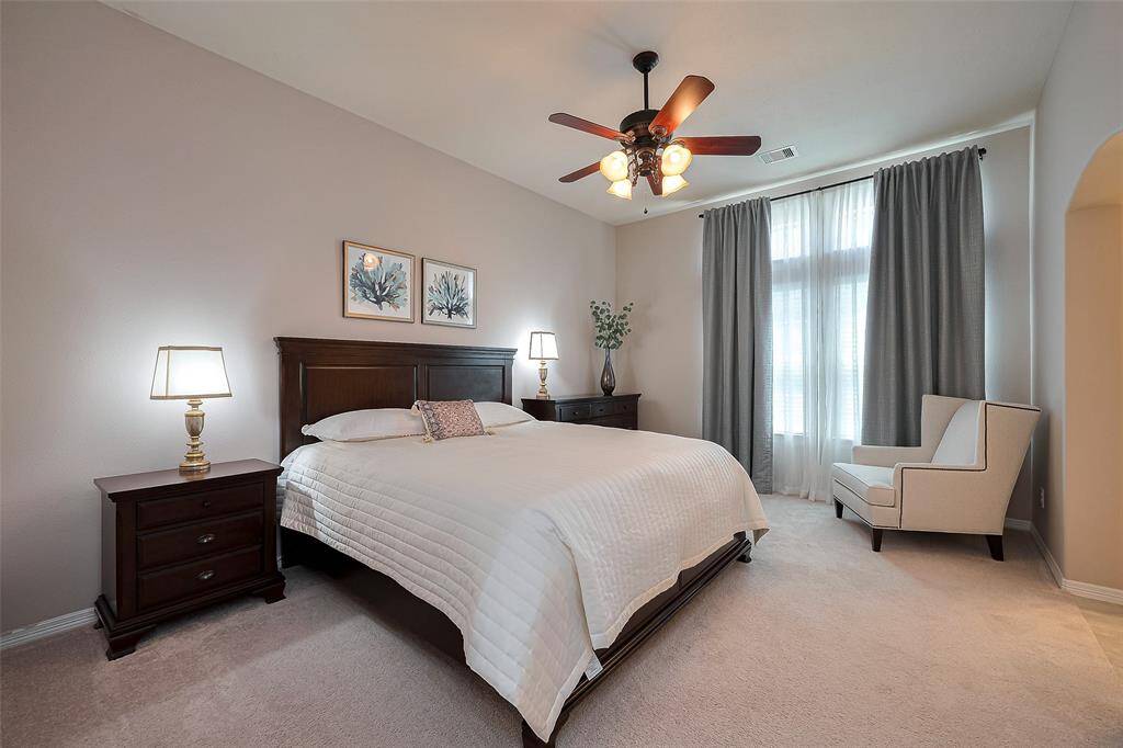 This is a cozy bedroom featuring a large bed with a wooden headboard, two bedside tables with lamps, and a comfortable armchair by a window with curtains. The room has soft carpeting, a ceiling fan, and is well-lit, creating a warm and inviting atmosphere.