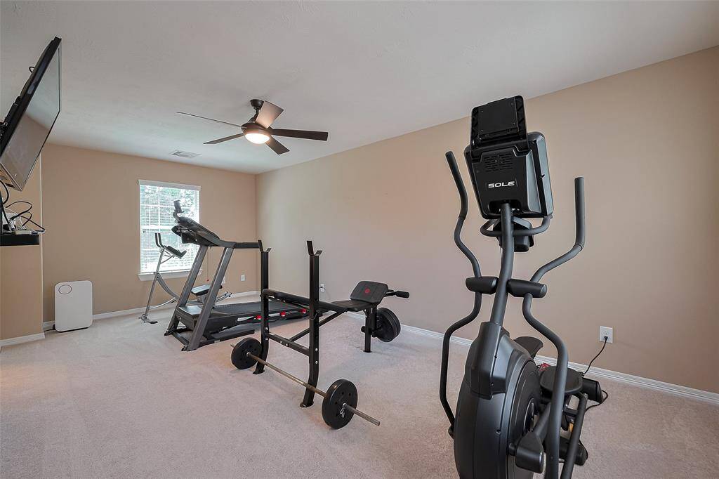 This bright and spacious room current used as a home gym features a treadmill, elliptical, and weight bench, with a ceiling fan and wall-mounted TV for entertainment and monitors security.
