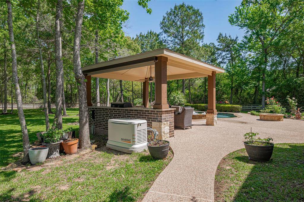 This photo showcases a serene backyard with a covered patio featuring comfortable seating, surrounded by lush greenery. A paved pathway leads to the patio area, and there's a Generac 26 kW generator visible, indicating preparedness and convenience. Ideal for outdoor relaxation and entertaining.