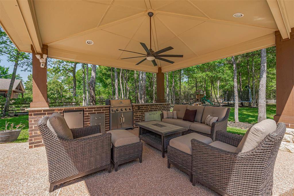 This photo showcases a beautiful outdoor patio area with comfortable wicker seating, a central coffee table, and a built-in grill under a covered pergola. The space is enhanced by a ceiling fan and surrounded by lush greenery, creating a perfect setting for relaxation and entertaining.