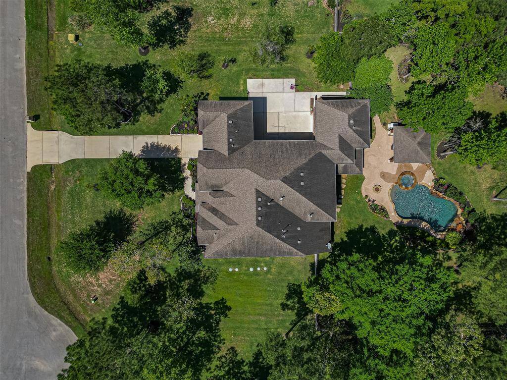 This aerial view showcases a spacious home with a well-maintained backyard, featuring a pool and surrounding patio. The property includes a long driveway, ample green space, and is bordered by mature trees, offering privacy and a serene environment.