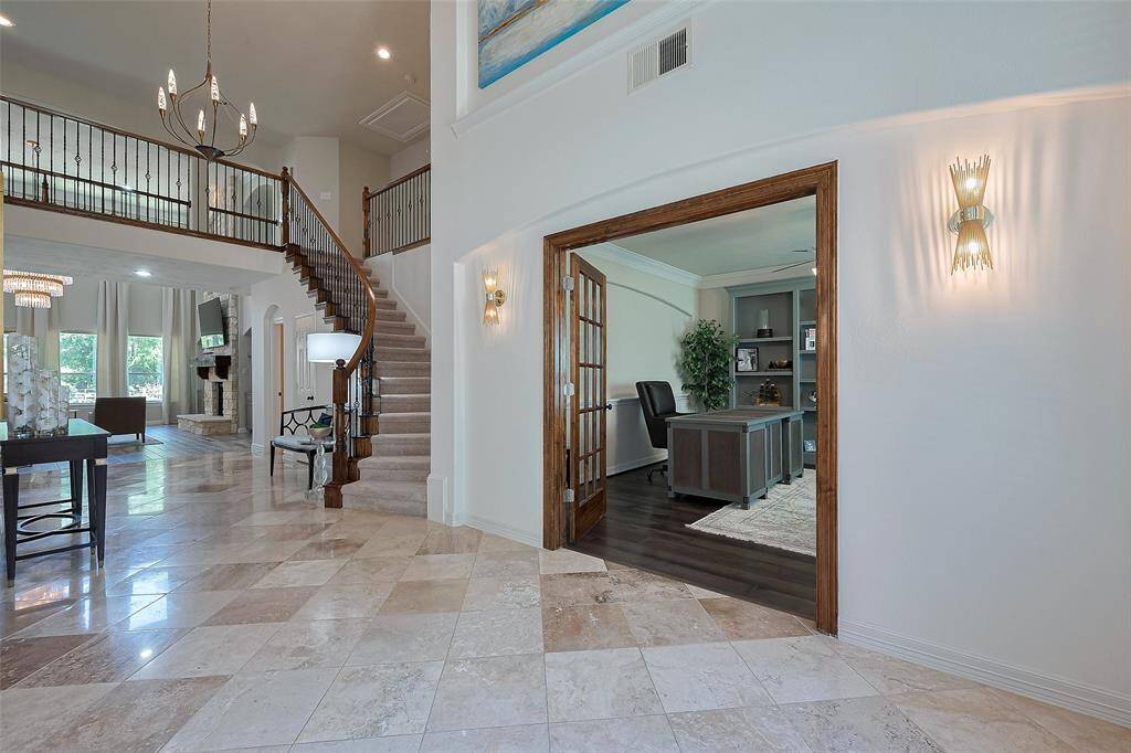 This home features a spacious, elegant foyer with polished tile flooring, a grand staircase, and modern lighting fixtures. The open layout includes a view into a cozy living area with large windows and a stone fireplace. There's also a welcoming office space with wooden floors and built-in shelving.