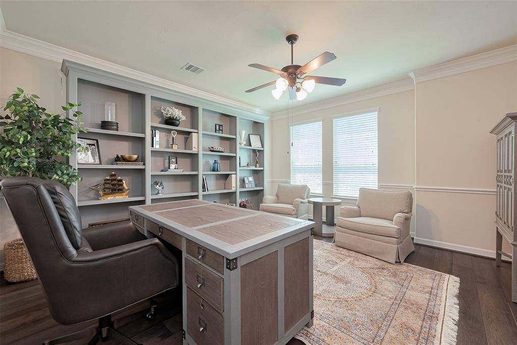 This photo showcases a bright, inviting home office with built-in shelves, a large desk, and comfortable seating. Natural light fills the room through two windows, and the space is accented with elegant crown molding and a ceiling fan. Flooring is laid with brand new wooded floors.