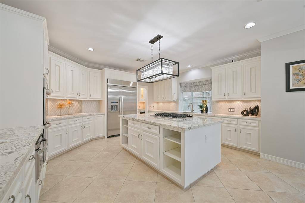 Another view of this modern Chef's kitchen featuring sleek, white cabinetry, under cabinet lighting, a large central island with a Viking cooktop, and elegant pendant lighting. Kitchen is connected to the Formal dining through the Butler's Pantry featuring glass inserted cabinet doors and a wine Fridge. Kitchen offers ample cabinet and counter space, and a bright, airy atmosphere filled with natural light from the windows.