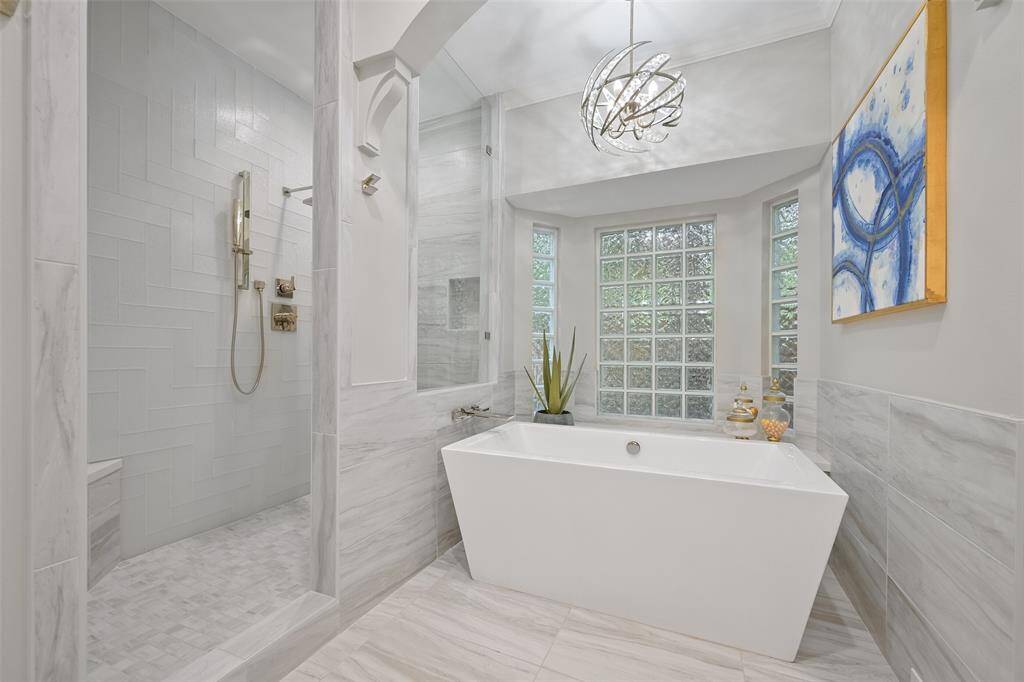 This elegant, designer-curated primary bathroom showcases a modern aesthetic with a freestanding soaking tub, spacious walk-in shower, and frosted glass block windows that offer both privacy and natural light. Thoughtfully selected fixtures and contemporary lighting enhance the luxurious, spa-like atmosphere.
