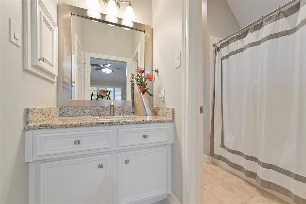 The first sink area of the Jack-n-Jill bathroom features a granite countertop with a stylish sink and sleek fixtures. It includes a large mirror with overhead lighting, white cabinetry, and a tub/shower combo. The neutral tones create a clean, inviting ambiance.