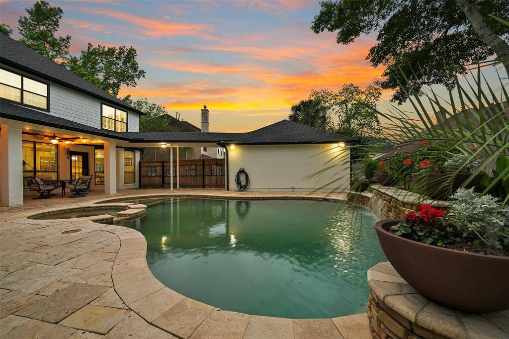 This inviting, tropical backyard features a sparkling pool/spa surrounded by travertine decking. The covered patio offers a relaxing seating area, perfect for outdoor dining. Lush greenery and vibrant flowers enhance the serene atmosphere, ideal for entertaining or unwinding at sunset.
