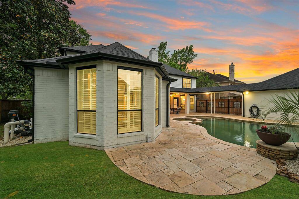 Another view of the beautifully landscaped backyard with additional green space, a curved pool surrounded by a stone patio. The unique, bay-windowed extension of the house overlooks the pool area. The ambiance is enhanced by a vibrant sunset sky, creating a serene and inviting outdoor space.
