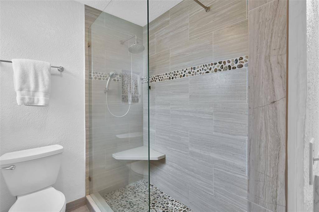 The Primary Bath continues with a frameless shower with rain showerhead and hand-held shower wand with temperature control located near the opening. Wood-look tile with pebble accents, product niche and sitting seat make showering a relaxing experience.