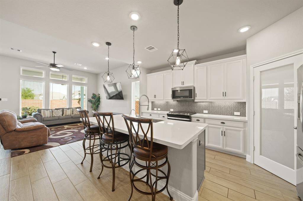Bright and open living space featuring a modern kitchen with a large island and bar seating. The area seamlessly connects to a cozy living room with ample natural light from large windows and stylish lighting fixtures.