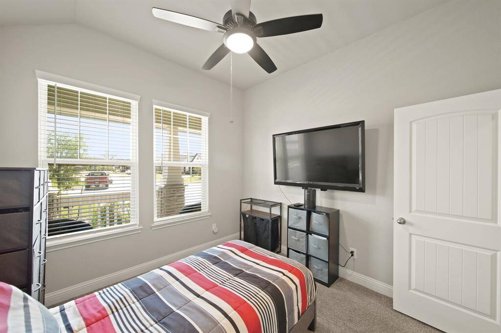 This bedroom features a large window with blinds, providing plenty of natural light. It includes a ceiling fan, a wall-mounted TV, and a neatly arranged storage unit. The space is carpeted and has a neutral color scheme, offering a cozy and practical setup.