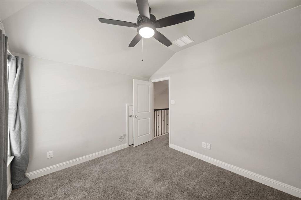This cozy room features neutral walls, soft carpeting, and a ceiling fan for comfort. A window with curtains allows natural light, and an open door leads to a hallway with a railing. Ideal as a bedroom or office space.