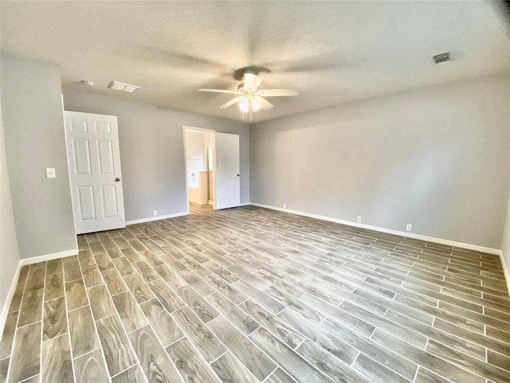 This spacious primary bedroom features modern wood-look tile flooring, neutral gray walls, and a ceiling fan. Perfect for a cozy living space or bedroom.