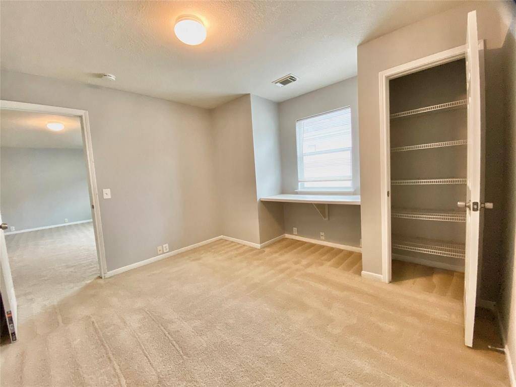 This room features soft carpeting, neutral walls, and ample natural light. It includes a built-in desk area under a window and a closet with shelving, making it perfect for a home office or study.