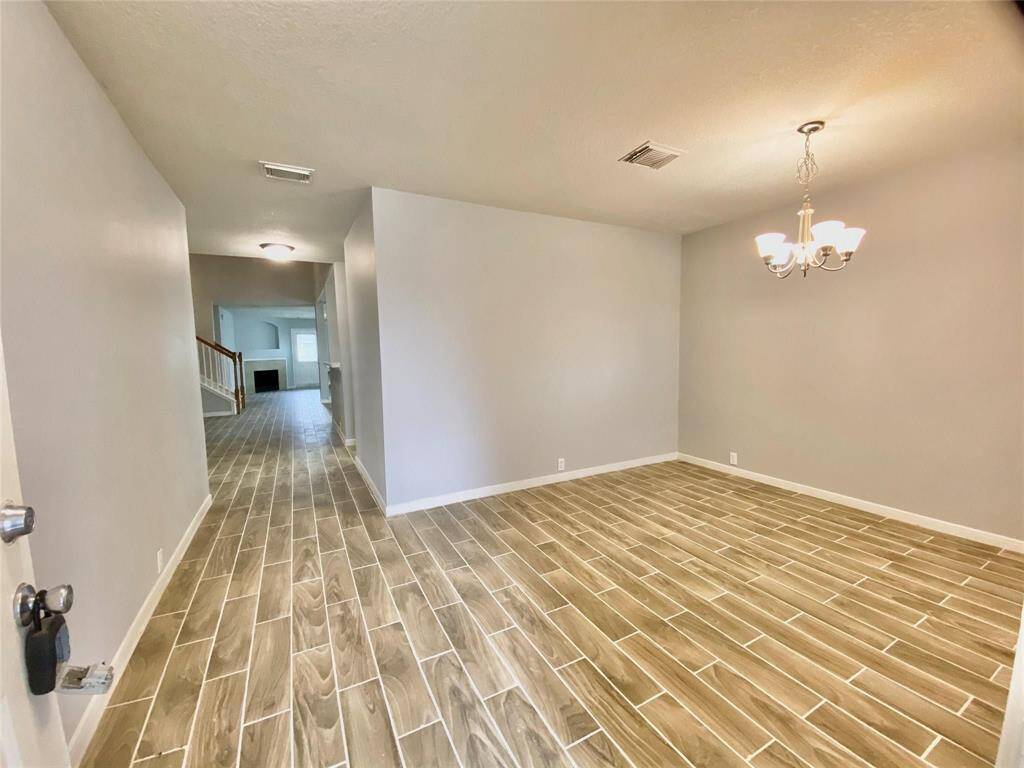 A spacious, open interior with wood-look tile flooring and neutral walls. A dining area features a modern chandelier, and the space leads to a living room with a visible staircase and fireplace.