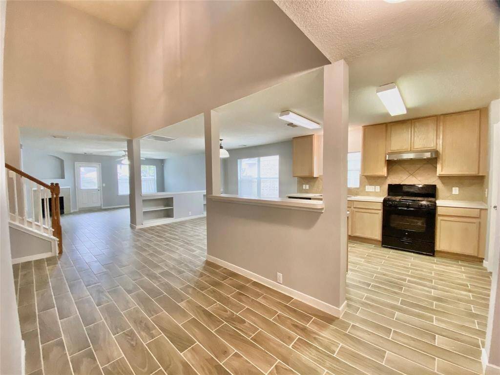 This photo showcases an open-concept living space with wood-like tile flooring. The kitchen features light wood cabinets and a black stove, with a breakfast bar connecting to the living area. Natural light fills the space, and there's a staircase leading to the upper level.