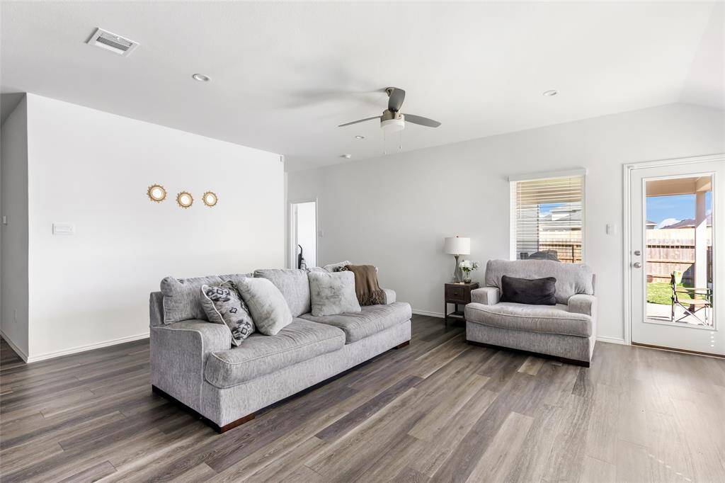 Bright, spacious living room with modern grey flooring and neutral walls. Features large windows providing natural light and access to the backyard through a glass door.