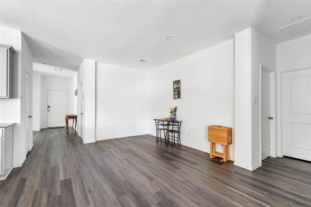 This photo shows a bright, open living space with modern wood flooring and white walls. There's a small dining area with a table and chairs, and it leads to a hallway with a visible entry door.
