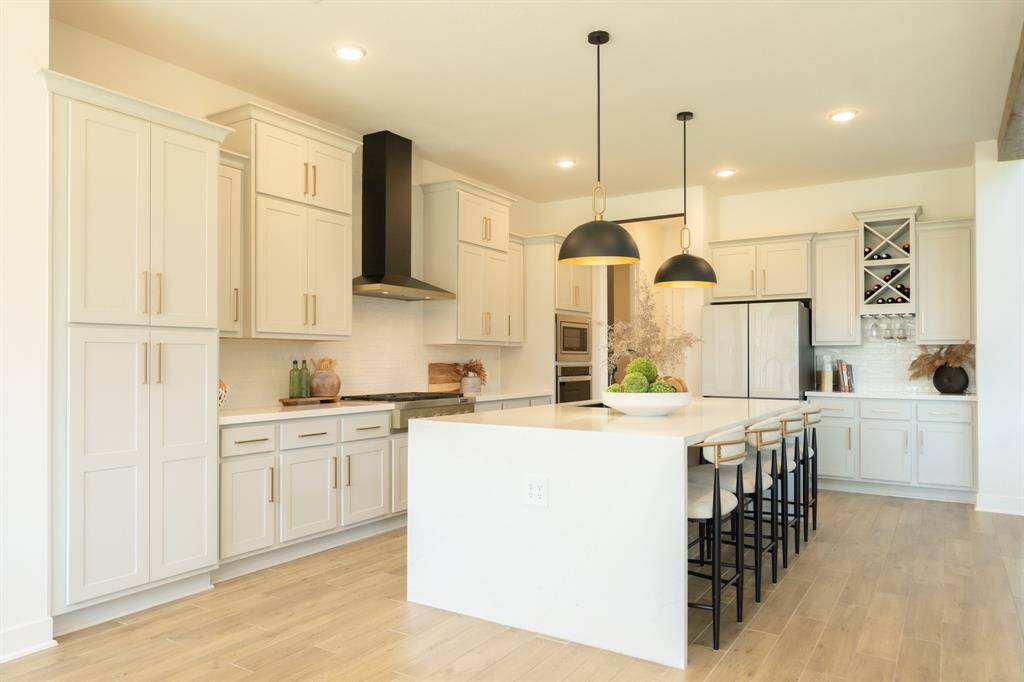 This elegant kitchen features sleek, soft gray cabinetry, a large island with seating, and modern black pendant lighting, creating a sophisticated and functional space perfect for both cooking and entertaining.  The light and airy design is complemented by warm, wood-look tile flooring