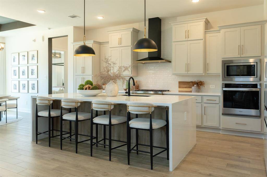 This bright, modern kitchen combines light cabinetry with white quartz countertops including the waterfall edge island, and sleek black and gold pendant lights, creating a refined, contemporary feel. Thoughtful styling and warm floors complete the elegant, open-concept space. Designer Detail - custom wrapped vent hood and soft brass cabinet hardware