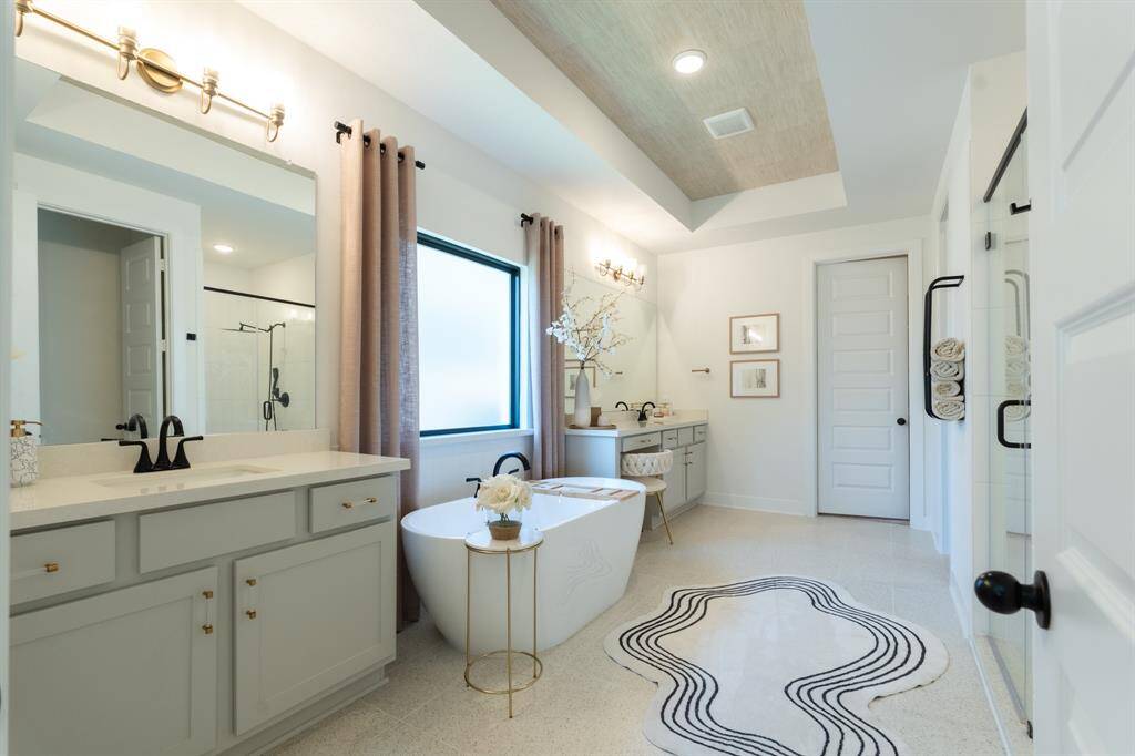This spa-like primary suite boasts a freestanding soaking tub, dual vanities with sleek gray cabinets and gold accents, and a walk-in shower, creating a luxurious and tranquil retreat.  The neutral color palette and natural light enhance the calming atmosphere. Designer details - custom painted window, towel rack, draperies and custom cabinet hardware