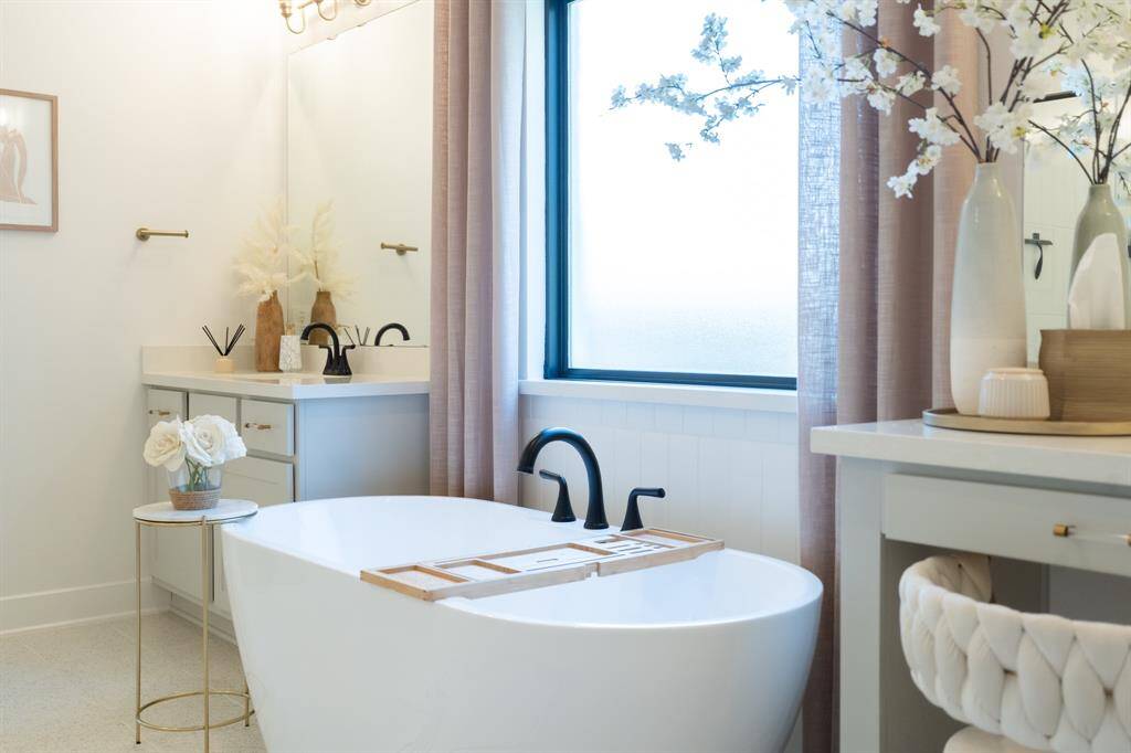 This luxurious primary bathroom features a freestanding soaking tub, a double vanity with sleek gray cabinets, and a large window allowing ample natural light. The neutral color palette and elegant fixtures create a serene and spa-like atmosphere.