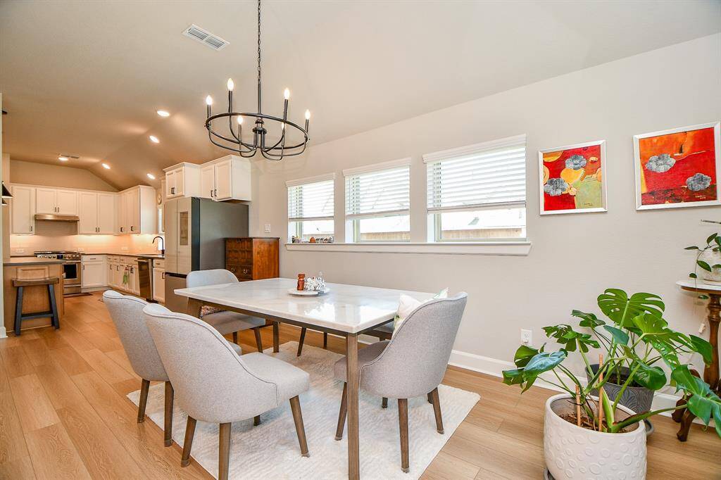 This photo showcases a bright, open-concept kitchen and dining area with light wood flooring. The kitchen features white cabinetry, modern appliances, and a central island. The dining space is adorned with a sleek table, upholstered chairs, and a stylish chandelier, complemented by natural light from multiple windows.
