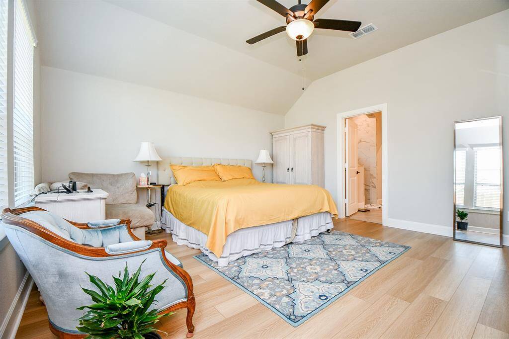Bright and spacious bedroom with hardwood floors and vaulted ceiling. Features a cozy bed with yellow bedding, a stylish armchair, and a full-length mirror. Large windows provide ample natural light. Connected en-suite bathroom with modern fixtures.