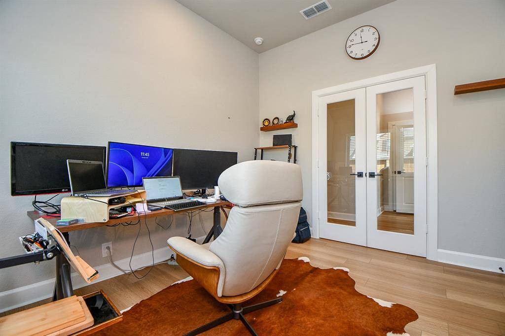 This home office features a modern setup with multiple monitors on a spacious desk, a comfortable chair, and light wood flooring. It includes glass double doors for a bright, open feel, and minimalist decor with a wall clock and shelving.