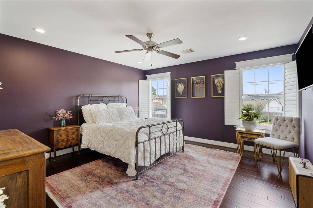 This secondary bedroom is a serene retreat with rich hardwood floors, deep plum accent walls, and large windows that flood the space with natural light. Custom shutters offer privacy while maintaining a bright and airy feel.