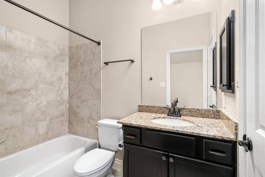 The secondary bathroom is designed with neutral tones and high-end finishes. It includes a granite-topped vanity, an oversized mirror, and a tiled tub/shower combo with sleek bronze fixtures.