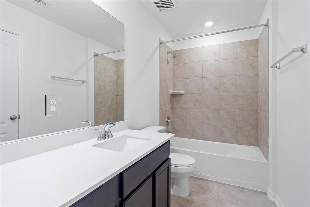 Secondary bath features tile flooring, bath/shower combo with tile surround, stained wood cabinets, beautiful light countertops, mirror, dark, sleek fixtures and modern finishes.
