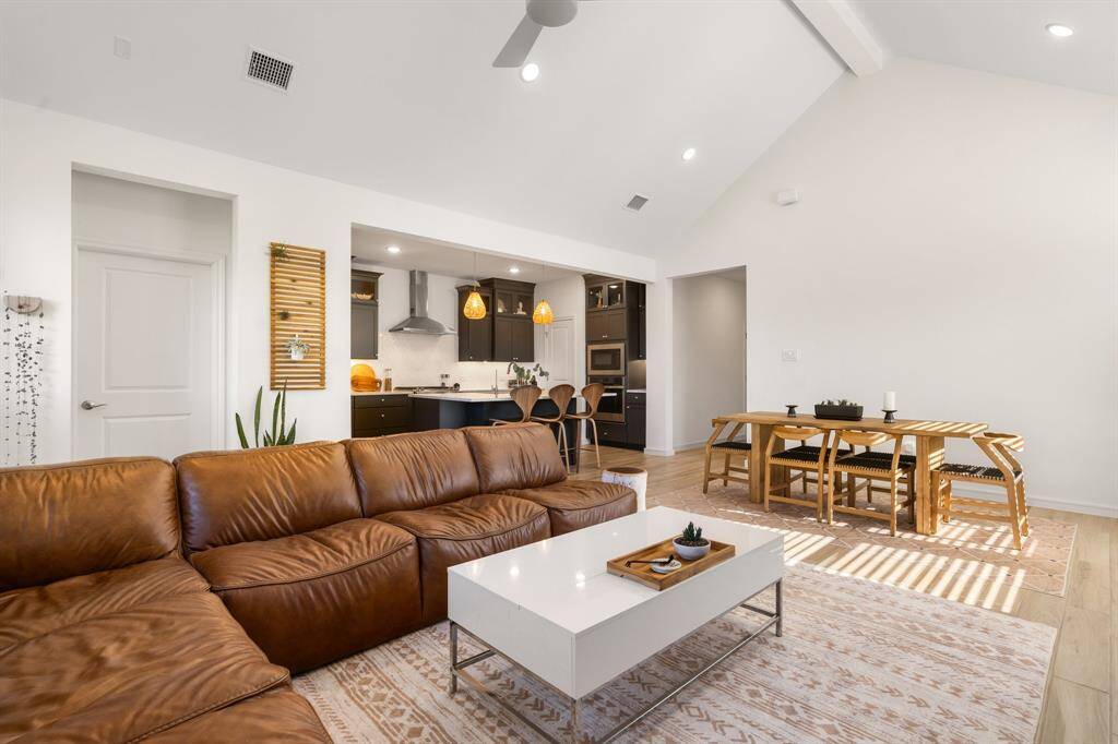 This photo showcases a spacious, open-concept living area with high ceilings and abundant natural light. It features a cozy brown leather sectional, a modern kitchen with pendant lighting, and a dining area with a wooden table. The space has a warm, inviting atmosphere with light wood flooring and minimalist decor.