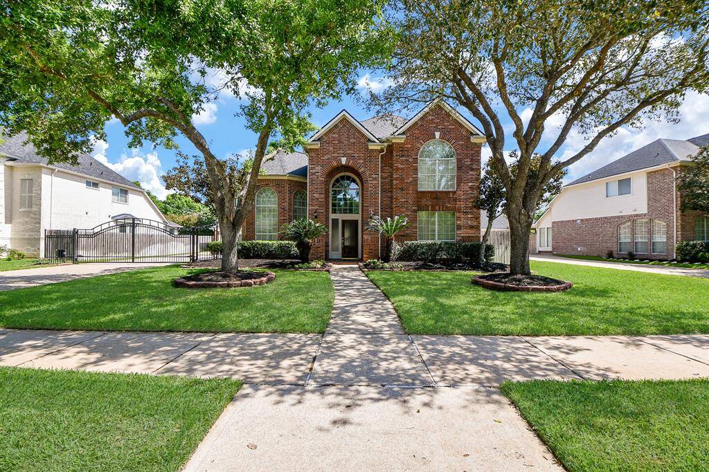 This home features a classic brick exterior with large arched windows and a welcoming double-door entrance. It's framed by mature trees and a well-maintained lawn, offering curb appeal in a suburban neighborhood.