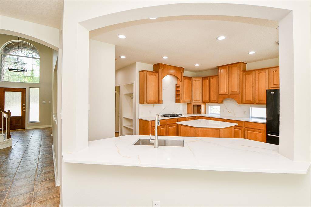This photo shows a modern kitchen with wooden cabinetry, a spacious quartz countertop, and built-in appliances. There's an open layout with an archway leading to a bright entryway featuring large windows.