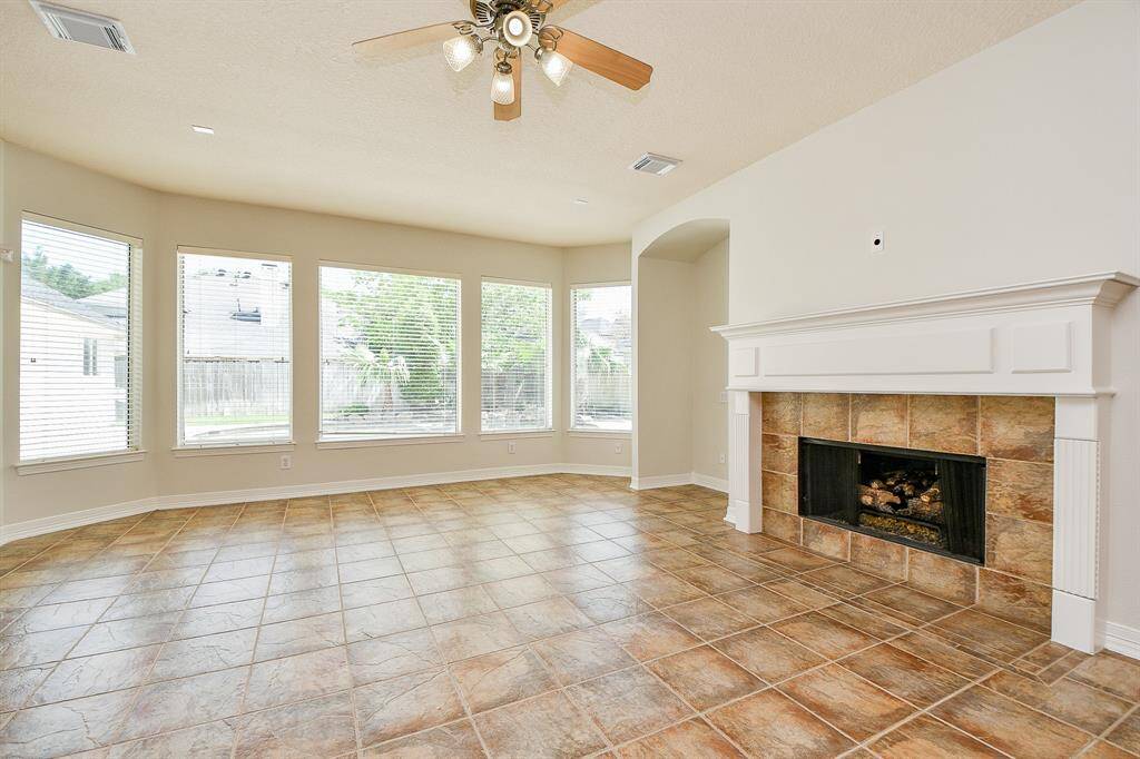 This spacious living room features large windows for natural light, a tiled floor, a cozy fireplace with a mantel, and a ceiling fan, creating a welcoming and bright atmosphere.