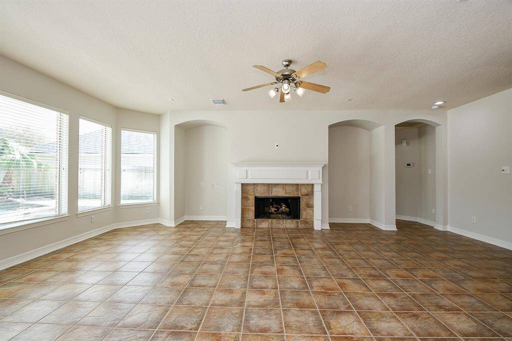 Bright living room with large windows, neutral walls, central fireplace with a white mantle and ceiling fan. Spacious and inviting.