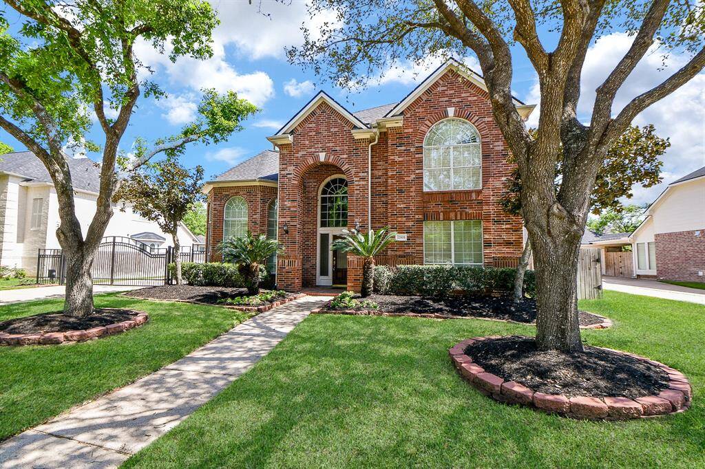 Charming two-story brick home with large arched windows, nestled between mature trees. The manicured lawn and landscaped garden beds frame a welcoming entrance, creating great curb appeal.