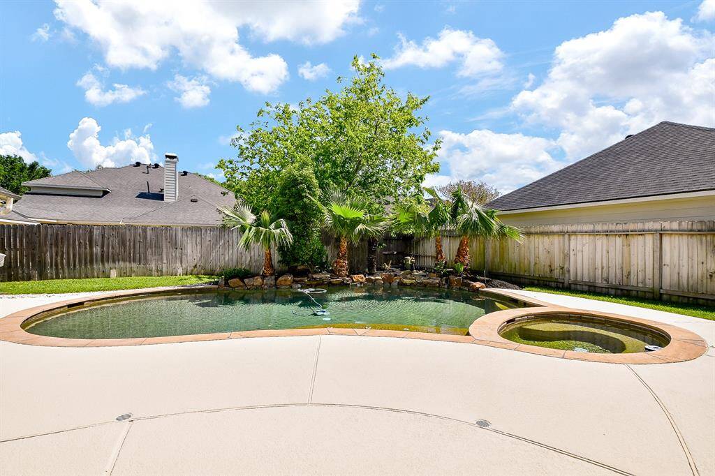 This backyard features a beautiful pool with a connected spa, surrounded by a spacious patio. Lush greenery and palm trees provide a tropical feel, while a wooden fence offers privacy.