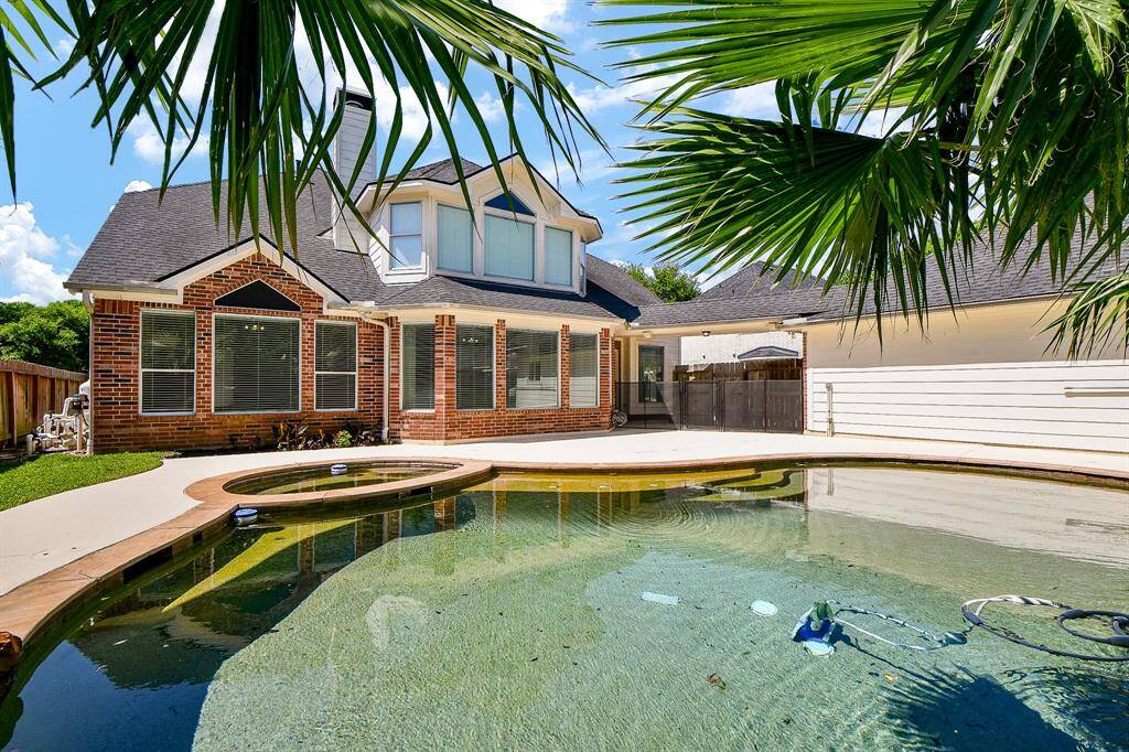 This inviting home features a spacious backyard with a well-maintained pool, surrounded by a charming brick exterior. The area is partially shaded by palm trees, offering a serene and private outdoor retreat.
