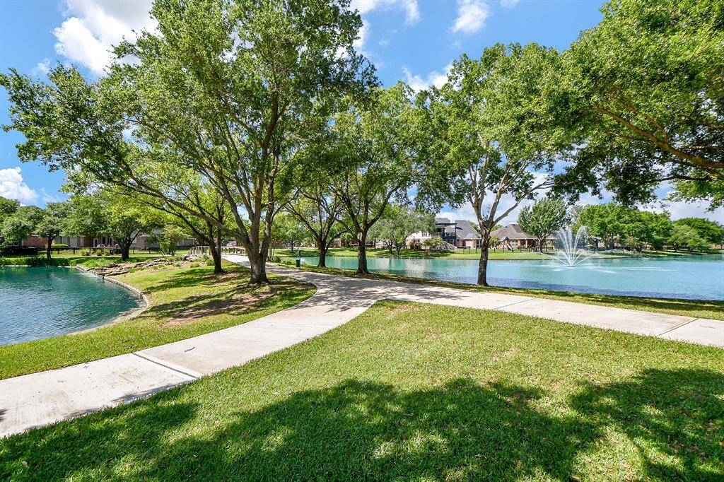 Serene view across from the house with a paved walkway, lush trees, and a lake featuring a fountain. Ideal for relaxation and outdoor activities in a peaceful neighborhood setting.