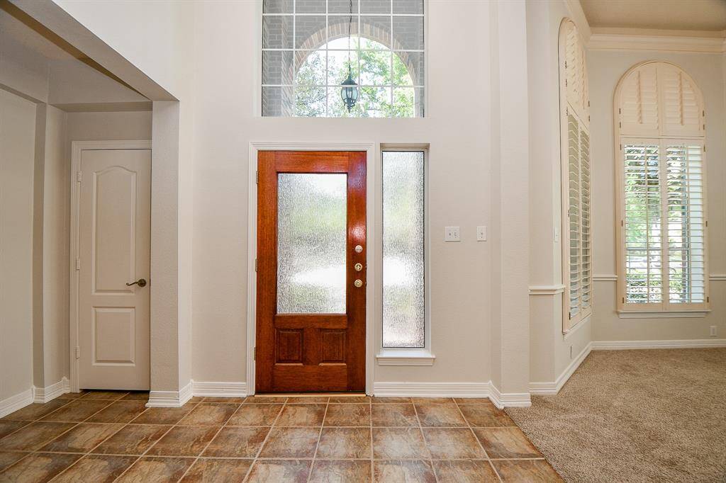 This entryway features a wooden front door with a frosted glass panel and a large arched window above, allowing natural light to brighten the space. The tiled floor provides durability, while the adjacent carpeted area offers warmth. Tall windows with shutters add elegance and privacy.