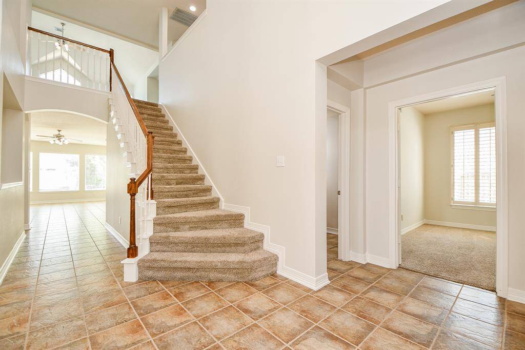 This photo shows a bright, open foyer with tiled flooring and a carpeted staircase. The space on the right leads to the study room with a large window.