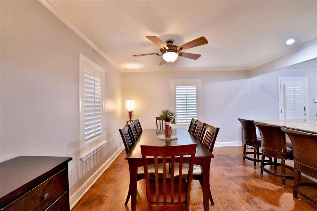 This photo showcases a spacious dining area with warm wood flooring, a central wooden dining table, and matching chairs. The room features a ceiling fan and ample natural light from windows with shutters. Adjacent is a kitchen counter with bar stools, creating an inviting open-plan layout.